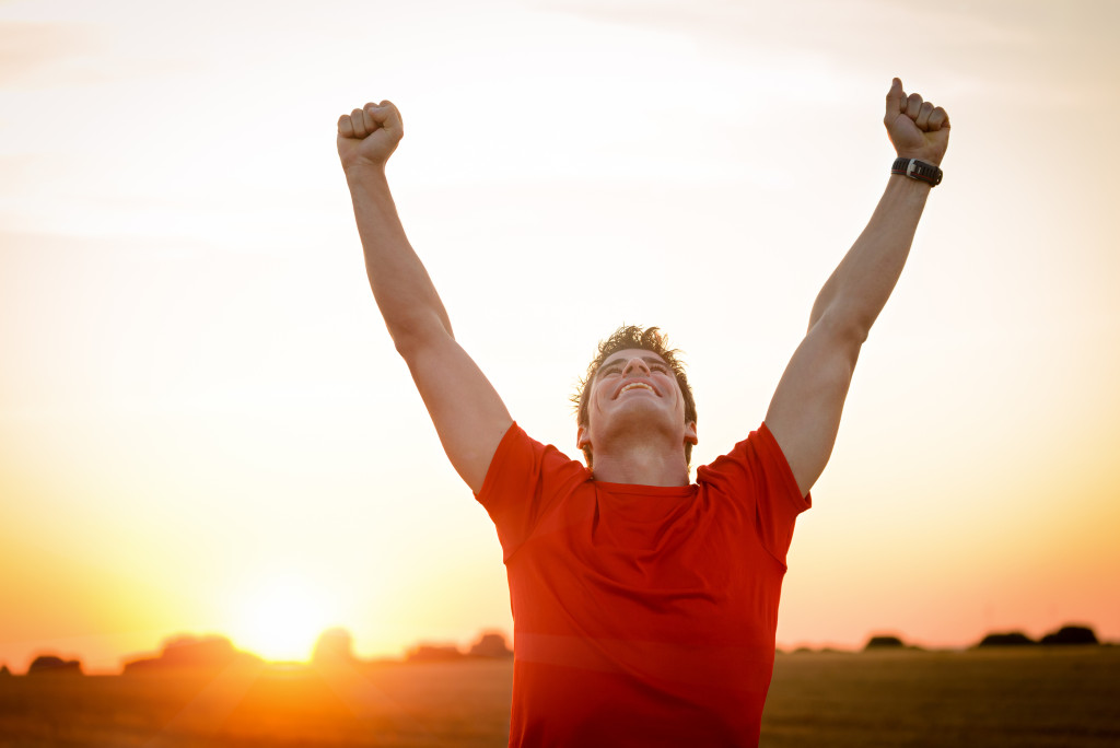 Successful man raising arms after cross track running on summer sunset. Fitness male athlete with arms up celebrating success and goals after sport exercising and working out.